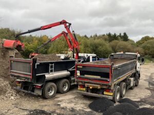 1 grabber lorry, on the left, facing the back, using its grabber to pick up material. Alongside the grabber's right is a tipper lorry, also facing the back.