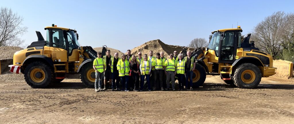 A team photo. 15 people, all wearing high visibility vests, standing in a row, between two yellow diggers, which are facing each other.