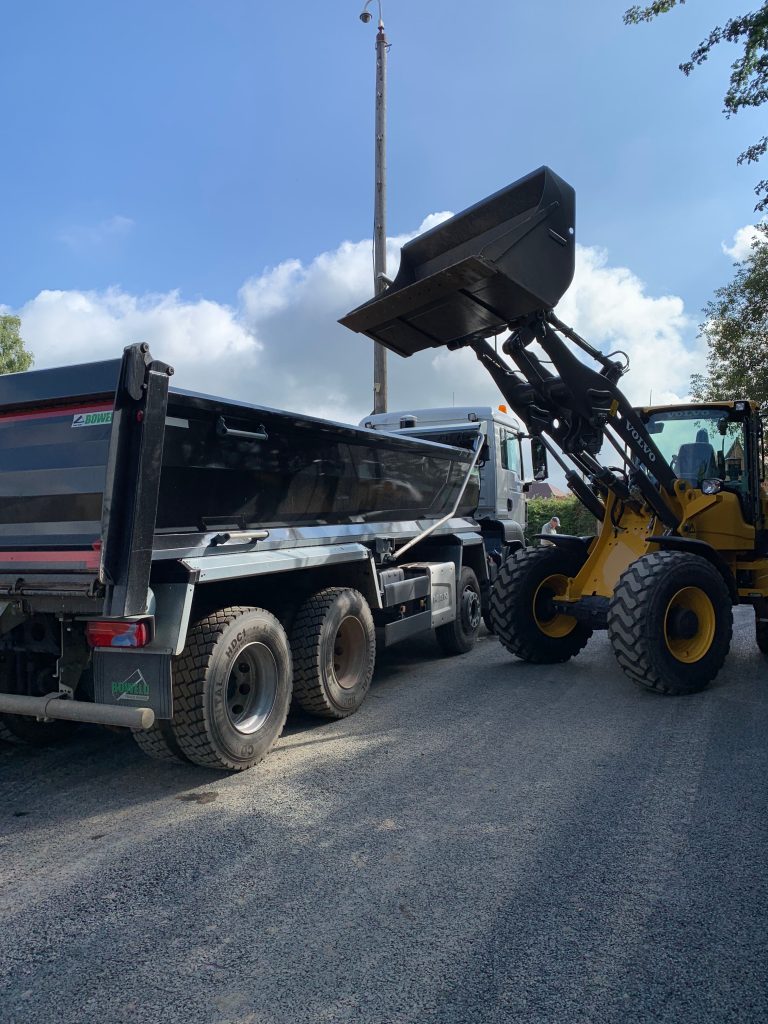 A tipper lorry, on the right, facing to the back, with a yellow digger, facing the lorry, with it's bucket raised high above the lorry's bed.