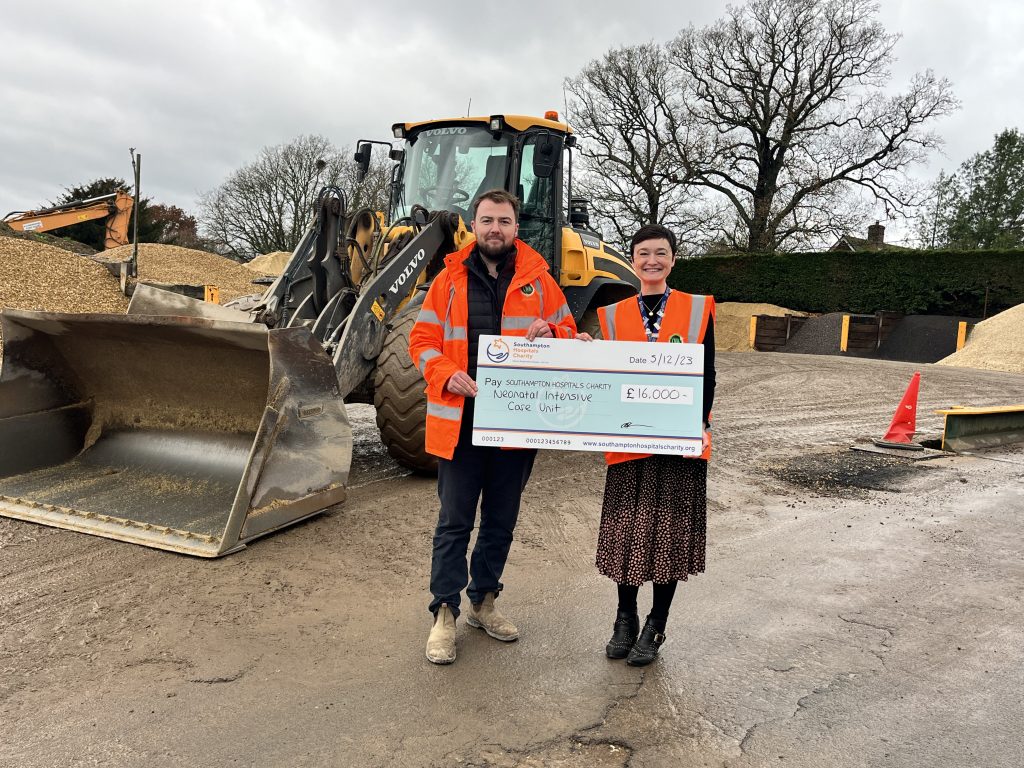 A man and a woman are smiling for the camera with an oversized cheque, to charity, for £16,000 They stand in the aggregate yard, in front of a large bucket, yellow digger.