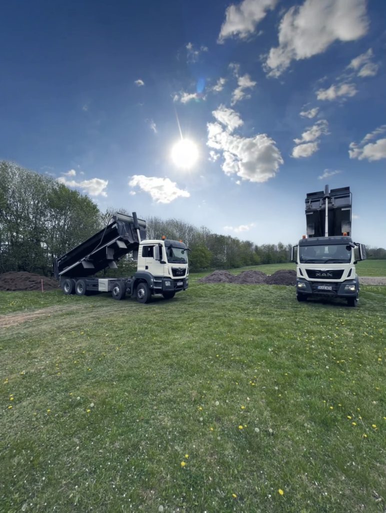 2 tipper lorries, dumping aggregate onto grassland. Both tipper beds are heightened to allow the aggregate to fall out. The sun is shining brightly in the middle of the sky