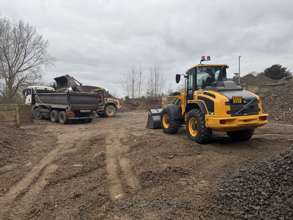 An aggregate yard, with 3 vehicles. A flat bed lorry, facing backwards, receiving aggregate, on its right hand side, from a yellow digger. here is another yellow digger in the forefront of the photo, facing towards the lorry and other digger.