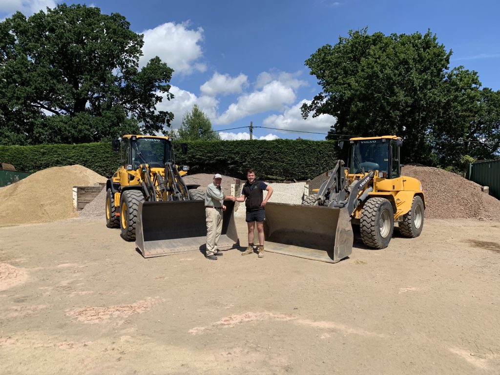 2 diggers facing towards each other, coming to a point, where 2 men (one wearing a baseball cap) are standing in front of each digger's bucket, shaking hands and smiling at the camera
