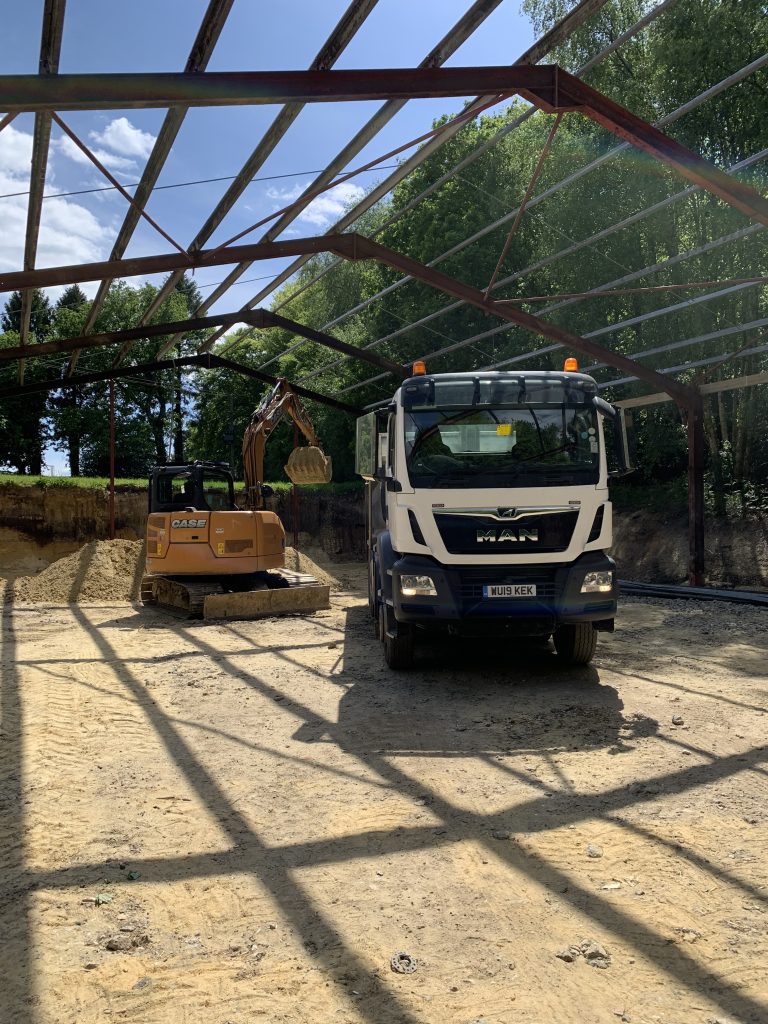 A yellow digger, on the left facing right, towards a tipper lorry facing forwards, in the dug out foundations of an open air structure. Rafters are in place of a roof